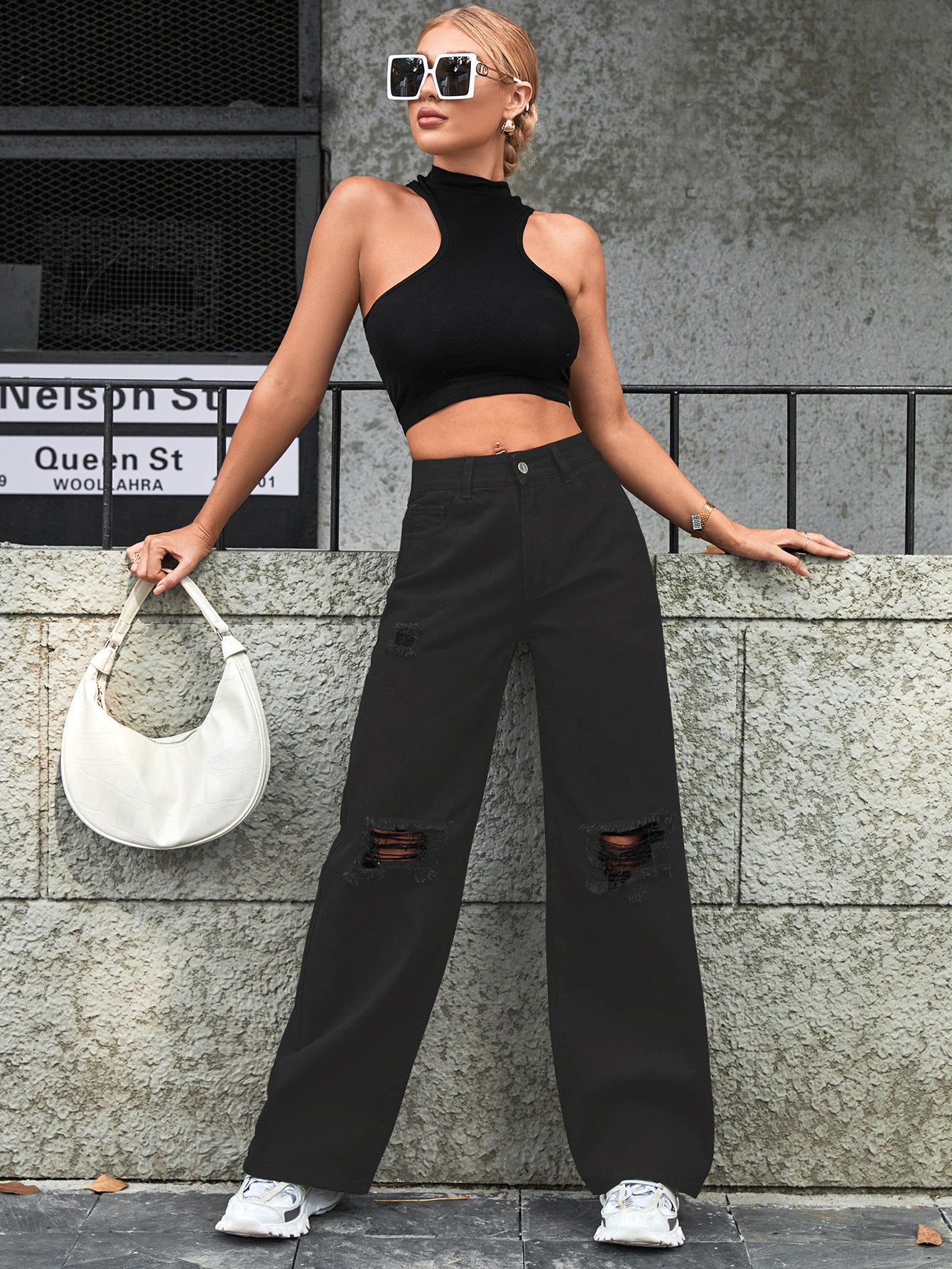 Women's high waist casual ripped jeans in black outfit with white bag against a stone wall