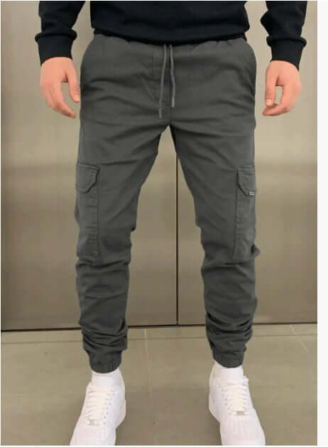 Man wearing gray baggy cargo jogger pants with patch pockets and white sneakers on a neutral background