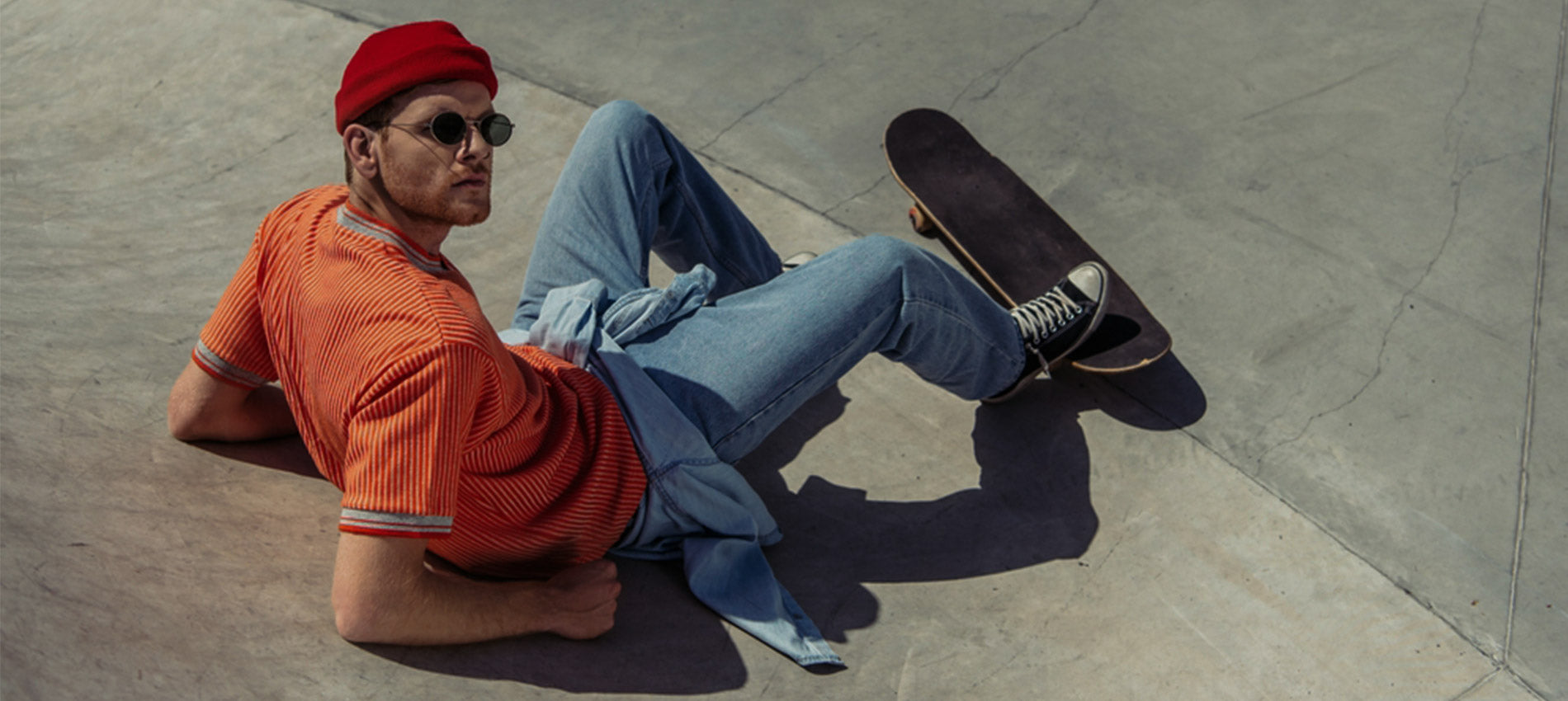 Person sitting on a concrete surface with a skateboard, wearing an orange shirt and blue jeans. - JeansSpot 