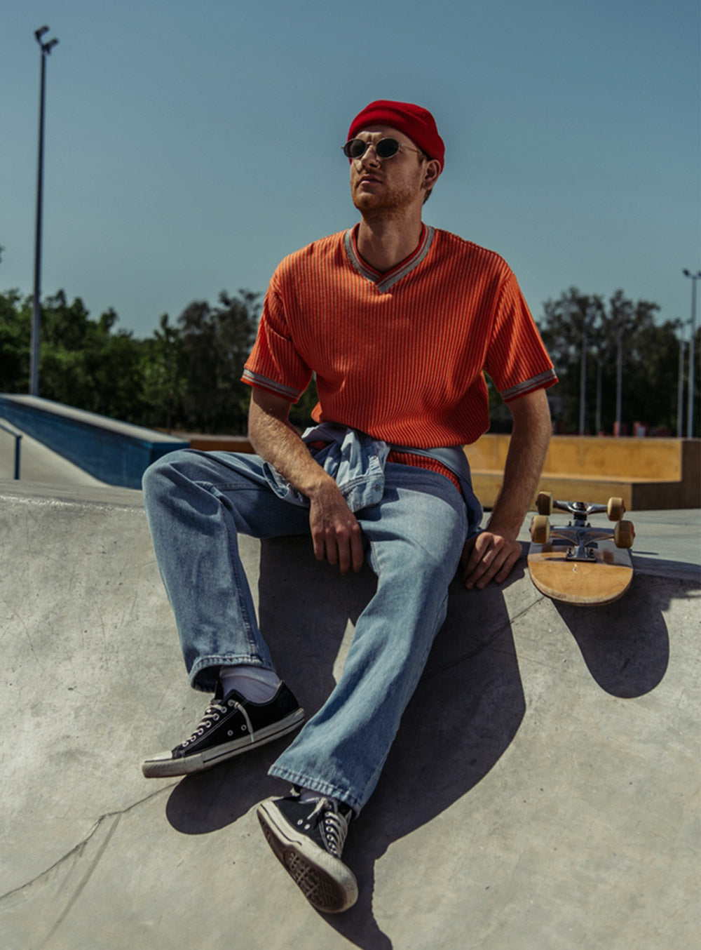 Person sitting on a concrete ledge with a skateboard, wearing an orange shirt and red cap. - JeansSpot 