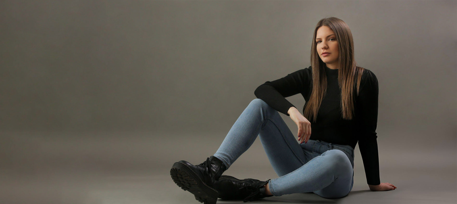 Woman sitting on the ground wearing black boots, light blue jeans, and a black top against a gray background - JeansSpot 