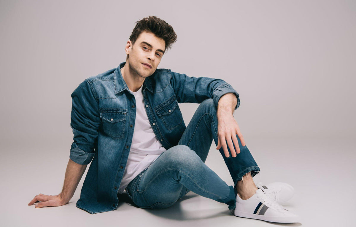 Man wearing a denim jacket, white t-shirt, jeans, and white sneakers sitting on a light gray background - JeansSpot 