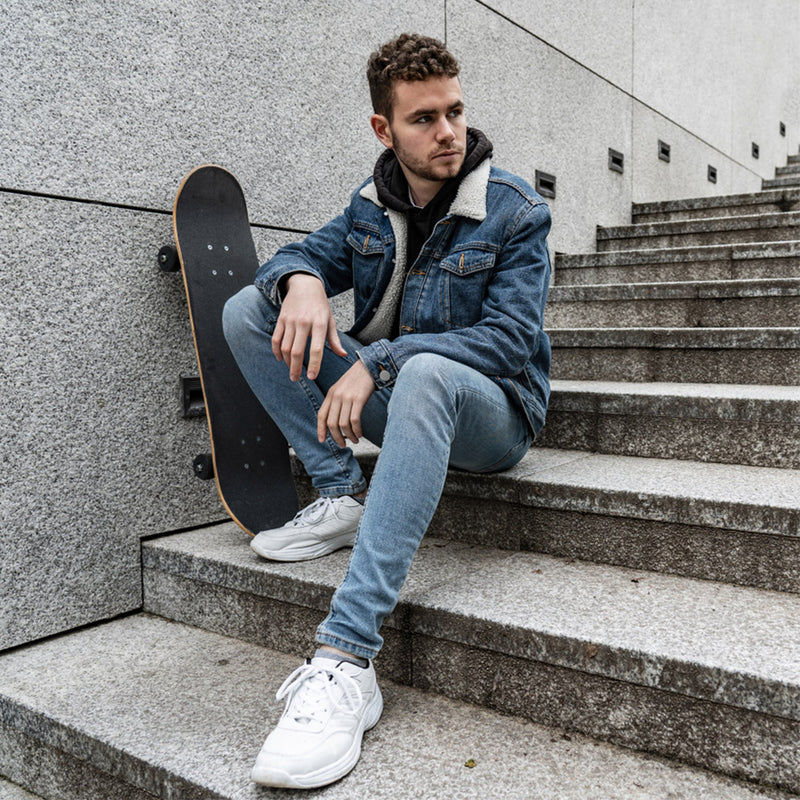 Man sitting on stairs with a skateboard, wearing a denim jacket and jeans. - JeansSpot 