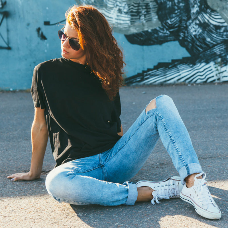 Woman sitting on the ground wearing a black t-shirt, blue jeans, and white sneakers with a graffiti wall in the background. - JeansSpot 