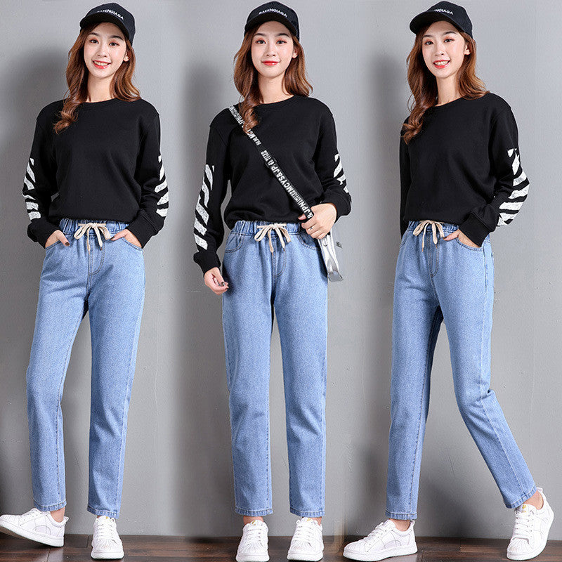 Woman wearing light blue high-waist jeans with a black sweater featuring white stripes and white sneakers on a gray background, casual denim outfit
