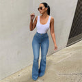 Woman wearing a white tank top and blue jeans standing against a gray wall.